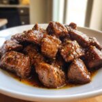 Close-up of tender, saucy crockpot steak bites piled high on a white plate.