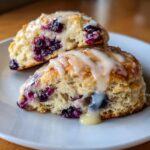 Two halves of flaky blueberry scones stacked, covered in a thick vanilla glaze dripping down the sides.