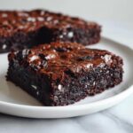 A close-up of a fudgy, dark chocolate square of oreo brownies with a shiny top, served on a white plate.