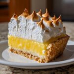 A thick slice of lemon meringue pie showing a graham cracker crust, bright yellow filling, and tall, torched meringue peaks.