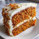 Close-up of a moist slice of carrot cake layered with cream cheese frosting and topped with nuts.