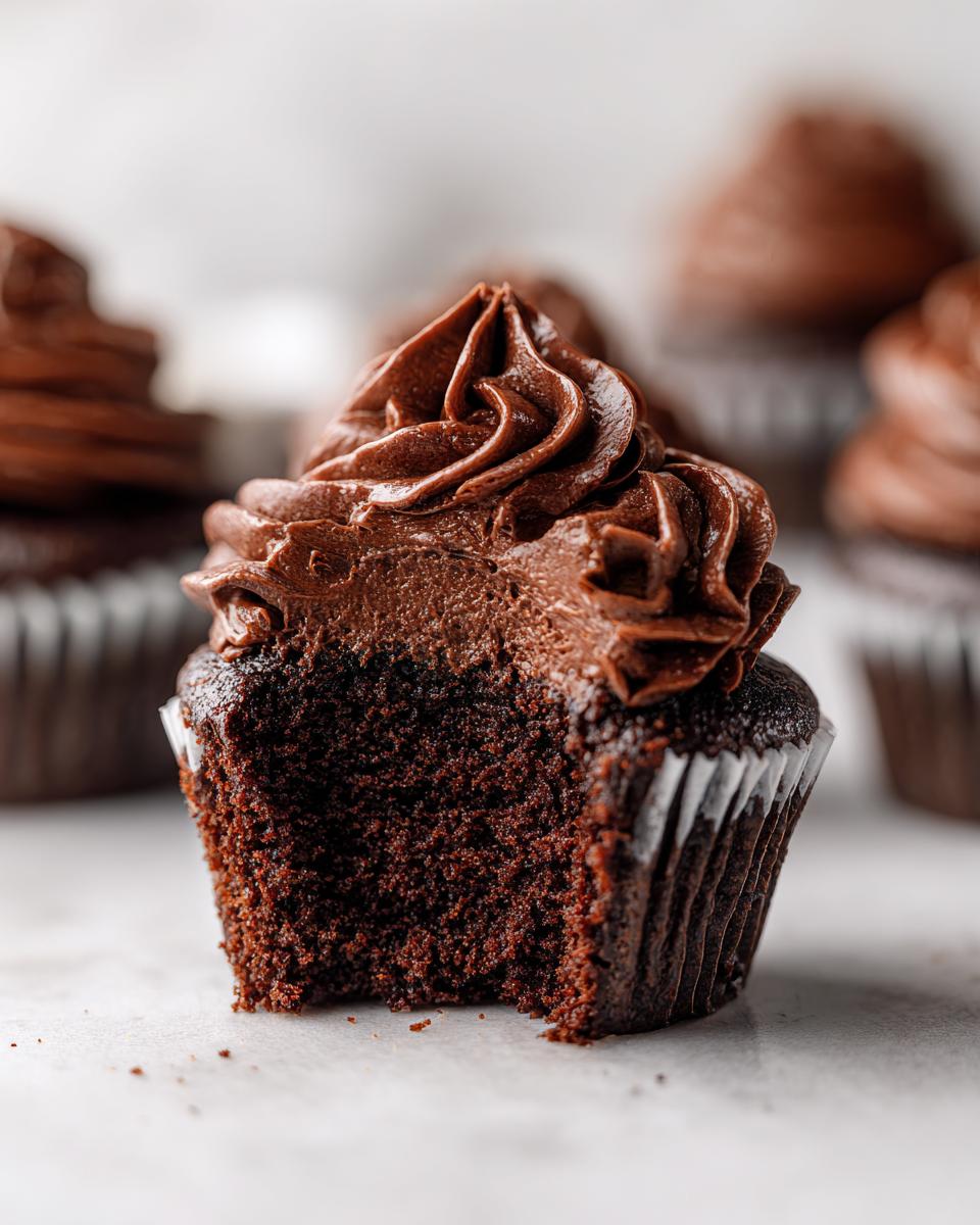 Close-up of a moist chocolate cupcake with a bite taken out, topped with rich chocolate frosting swirls.
