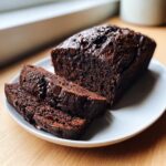 A moist, dark chocolate zucchini bread loaf with chocolate chips, partially sliced on a white plate.