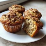 Four golden brown oatmeal muffins on a white plate, one broken open to show the moist interior.