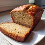 A loaf of golden brown protein banana bread with one thick slice cut and resting in front.