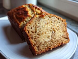 A close-up of a loaf of protein banana bread with one thick slice cut and leaning against the main loaf on a white plate.
