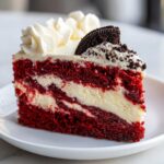 A decadent slice of red velvet cheesecake featuring alternating layers of red cake and creamy cheesecake, topped with white frosting and an Oreo cookie piece.