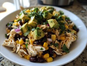A bowl of shredded chicken high protein salad topped with diced avocado, black beans, corn, and cilantro.