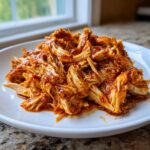 Close-up of shredded chicken tinga coated in a rich, red, spicy sauce piled on a white plate.
