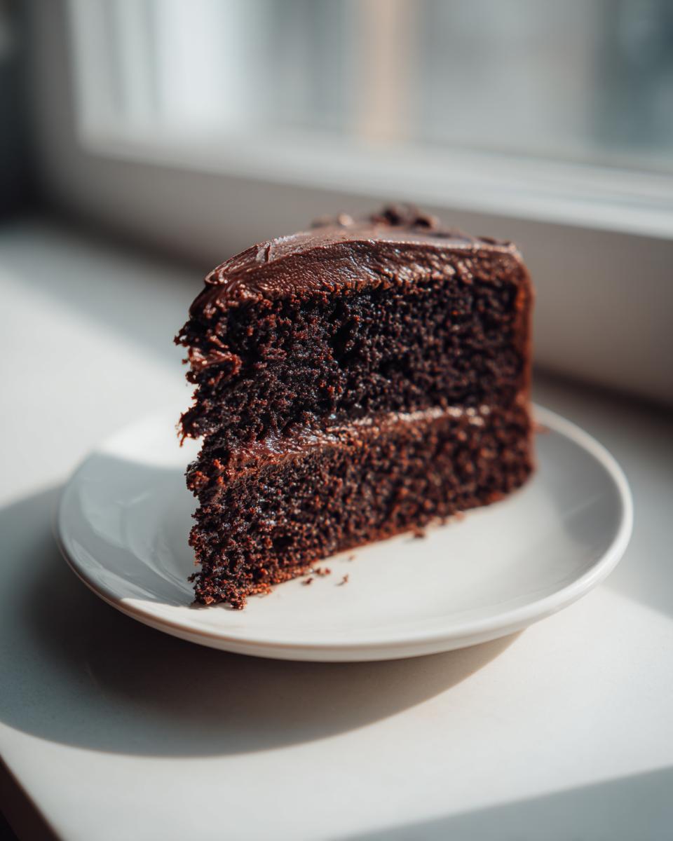 A moist slice of rich chocolate cake with dark frosting sitting on a white plate near a window.