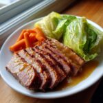 A plate featuring sliced, glazed corned beef served alongside boiled carrots and wedges of cabbage.