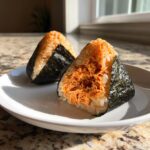 Two triangular tuna onigiri rice balls filled with spicy tuna mixture and wrapped with nori seaweed, sitting on a white plate.
