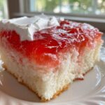 Close-up of a moist slice of jello poke cake featuring white cake soaked in red gelatin and topped with whipped cream.