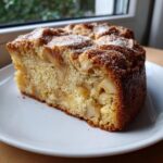 A thick slice of moist Swedish apple cake, studded with baked apple pieces and dusted with powdered sugar.