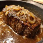 Close-up of a thick, seasoned pork steak simmering in a rich brown gravy topped with caramelized onions.