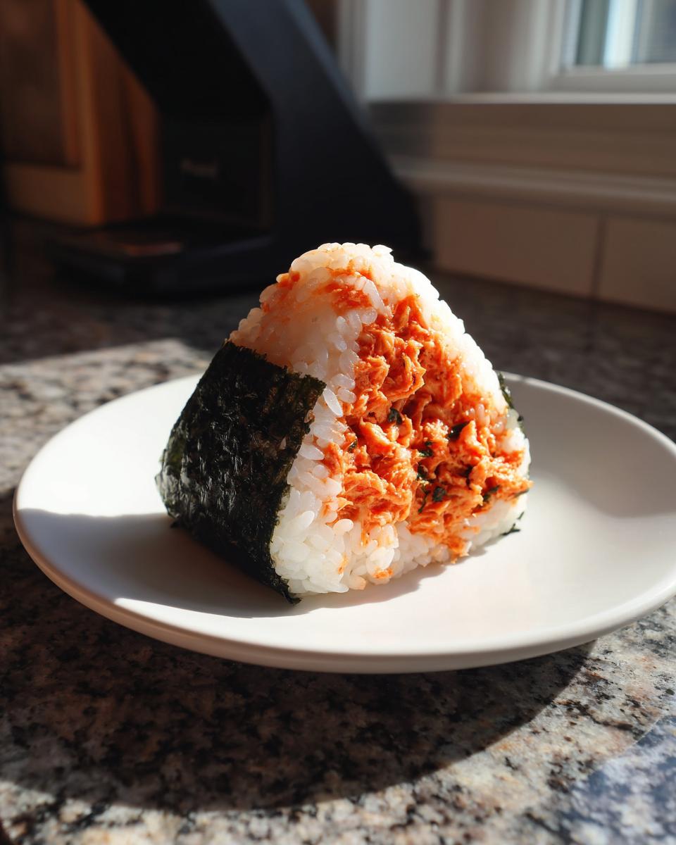 A single, triangular tuna onigiri filled with seasoned tuna and wrapped partially in dark nori seaweed, sitting on a white plate.