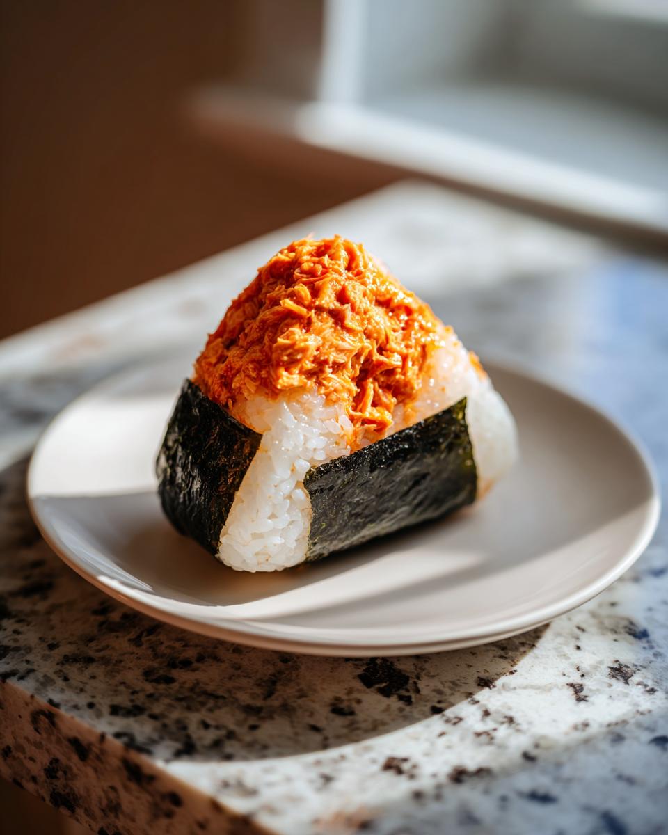 A single, triangular tuna onigiri wrapped in nori seaweed, topped with spicy orange tuna salad, sitting on a plate.