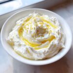 A close-up of freshly made whipped feta dip swirled in a white bowl, drizzled with olive oil and lemon zest.