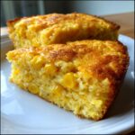 Two golden-brown slices of moist corn casserole recipe studded with whole corn kernels, served on a white plate.