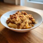 A close-up of a serving of amazing apple crumble in a white bowl, featuring soft baked apples and a crunchy, golden brown topping.