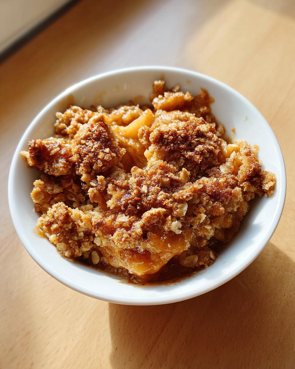 Close-up of a single serving of warm apple crumble with a crunchy topping in a white bowl.