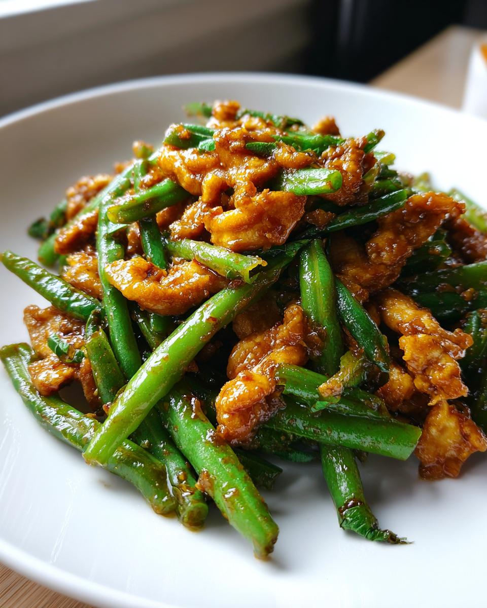A close-up serving of amazing chicken and green bean stir fry coated in a savory brown sauce on a white plate.
