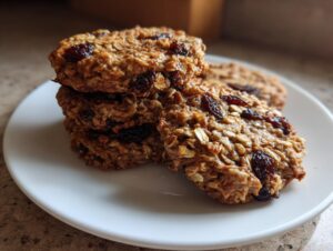 A close-up stack of four chewy oatmeal raisin cookies, rich brown in color, sitting on a white plate.
