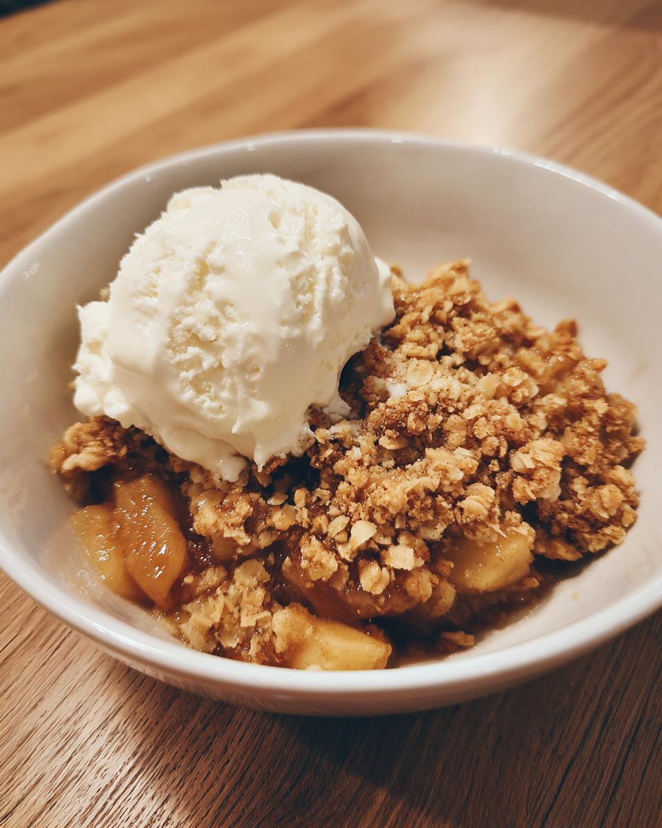 A close-up of a warm serving of apple crumble topped with a scoop of vanilla ice cream in a white bowl.