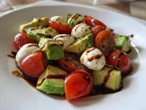 A close-up of a vibrant avocado caprese salad featuring chunks of avocado, cherry tomatoes, and mozzarella balls drizzled with balsamic glaze.