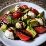 A vibrant close-up of avocado caprese salad featuring chunks of avocado, cherry tomatoes, mozzarella balls, and a balsamic glaze drizzle.