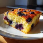 Close-up of a square slice of fluffy blueberry sheet pan pancakes on a white plate, bathed in sunlight.