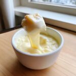 A piece of bread coated in thick, melted cheese being dipped into a small white bowl of cheese fondue recipe.