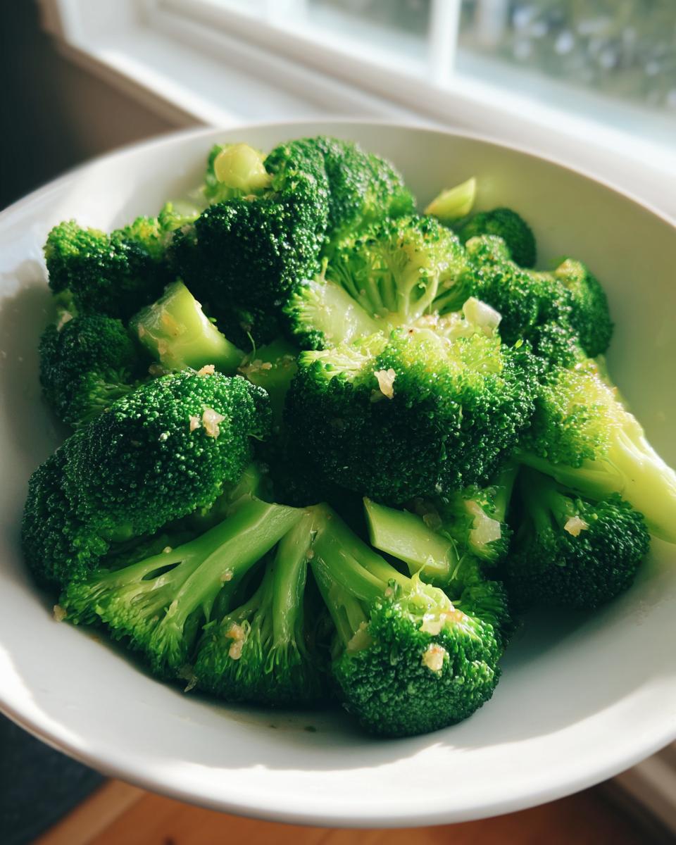 Close-up of bright green steamed broccoli florets lightly seasoned in a white bowl near a window.