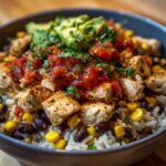A close-up, appetizing view of a chicken burrito bowl featuring seasoned chicken, rice, black beans, corn, salsa, and avocado.