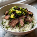 Close-up of a cilantro lime steak bowls layered with white rice, sliced steak, avocado chunks, and black beans.