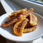 A pile of golden, crispy cinnamon sugar air fryer banana chips dusted heavily with sugar crystals on a white plate.