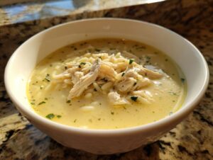 A close-up of a white bowl filled with Classic Greek Lemon Chicken Soup, featuring shredded chicken and orzo pasta.