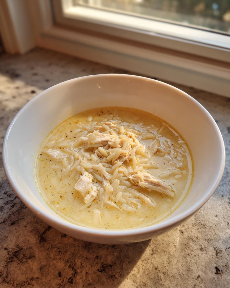 A white bowl filled with Classic Greek Lemon Chicken Soup, featuring shredded chicken and orzo pasta in a creamy yellow broth.