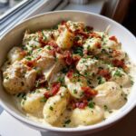 A close-up of a white bowl filled with Creamy Crack Chicken Gnocchi, topped with crispy bacon bits and fresh chives.
