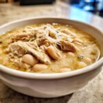 A close-up of a white bowl filled with creamy crockpot white chicken chili, featuring shredded chicken and white beans.