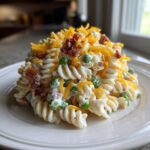 A mound of creamy ranch pasta salad featuring rotini pasta, green peas, bacon bits, and shredded cheddar cheese on a white plate.