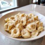A close-up of a bowl filled with creamy Tortellini Alfredo Recipe, topped generously with grated Parmesan cheese and black pepper.