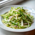 A mound of thinly sliced cucumber apple salad mixed with slivers of red onion in a light dressing.