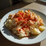 A close-up of a serving of creamy deviled egg potato salad generously topped with bright red paprika on a white plate.