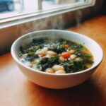 A steaming white bowl filled with Easy Tuscan White Bean Soup, featuring kale and carrots, set on a wooden table near a window.