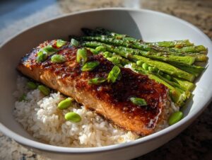 A perfectly cooked fillet of salmon with a thick, shiny honey glaze, served over white rice with asparagus and edamame in a white bowl.