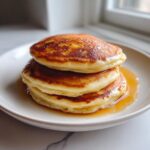 A close-up of three fluffy pancakes from scratch stacked on a white plate, drizzled with golden syrup.