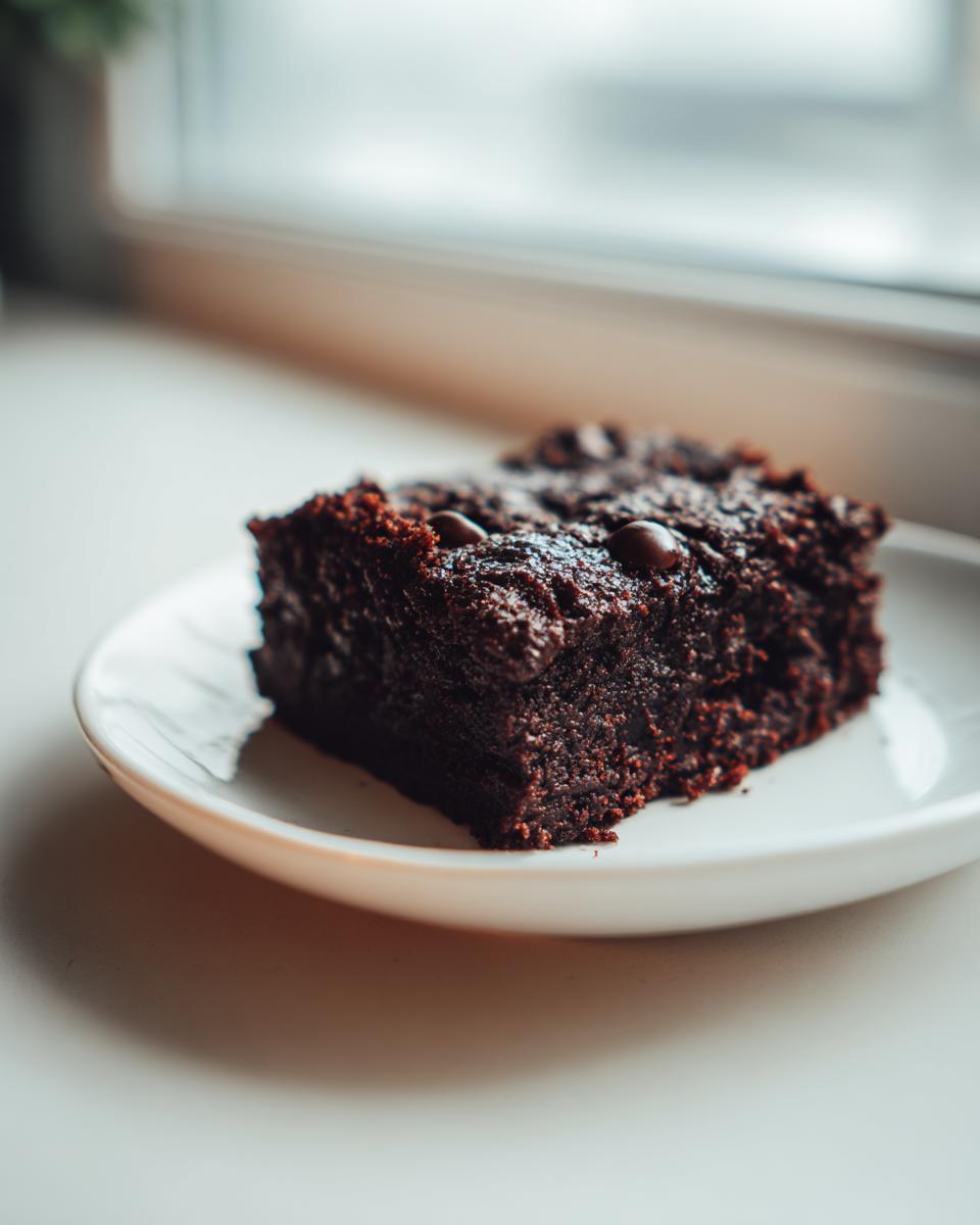 A close-up of a single, rich, fudgy slice of zucchini brownies topped with chocolate chips, resting on a white plate.
