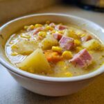 A close-up of a creamy bowl of ham potato corn chowder, featuring chunks of potato, ham, corn, and carrots.