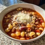 A close-up of a bowl of rich, tomato-based pasta fagioli soup with beans, ground meat, and pasta, topped with grated Parmesan cheese.